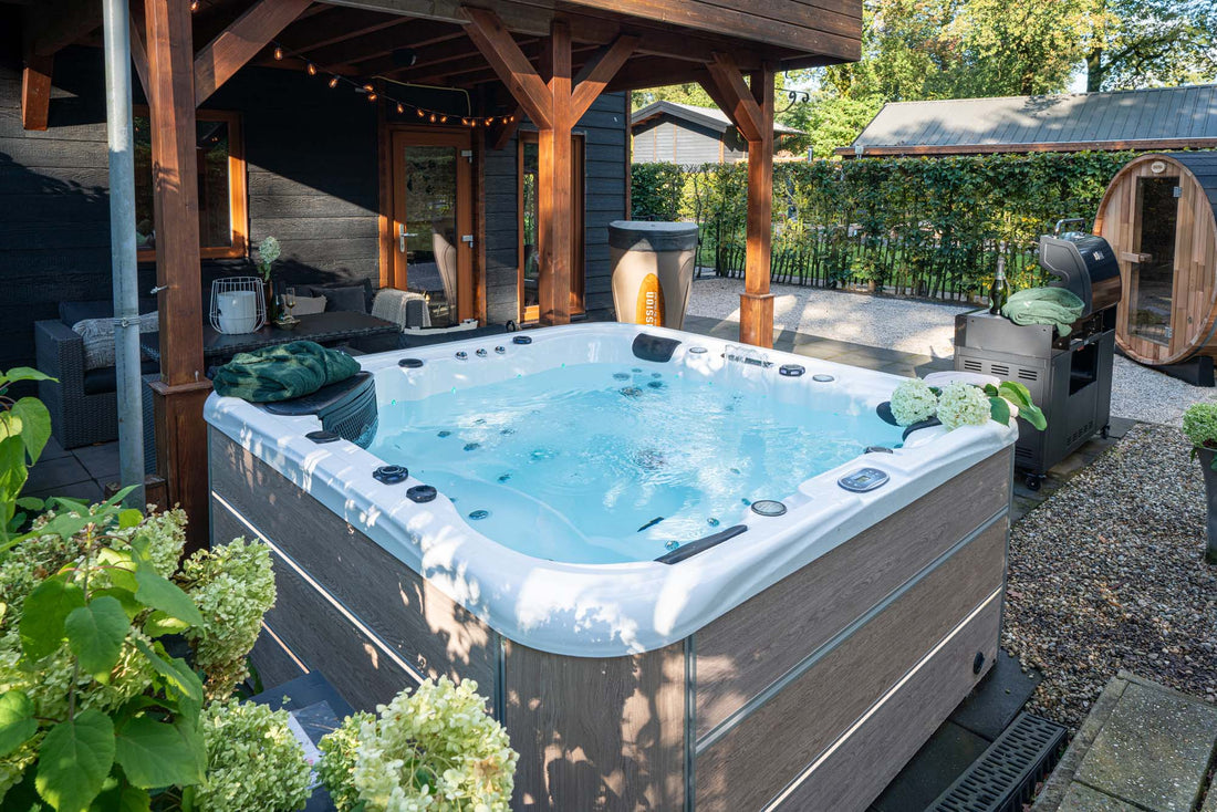 Een bubbelende jacuzzi op een terras naast een moderne bungalow, met buitenmeubilair, een barbecue en een barrelsauna op de achtergrond.